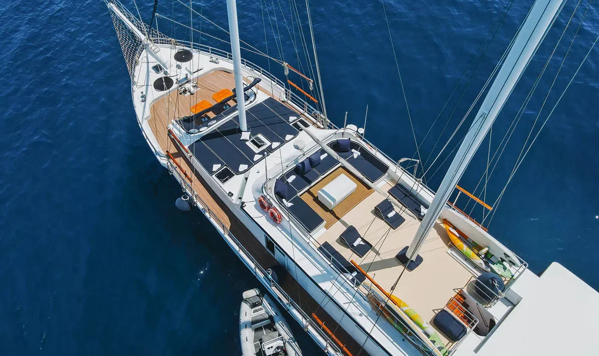 Sailboat viewed from above cruising on deep blue water, with a spacious wooden deck and seating areas visible on the deck.",