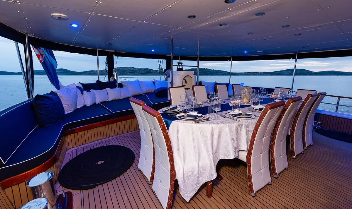 Luxurious dining area on a yacht with a long table set for dinner, white tablecloths, and blue evening lighting.