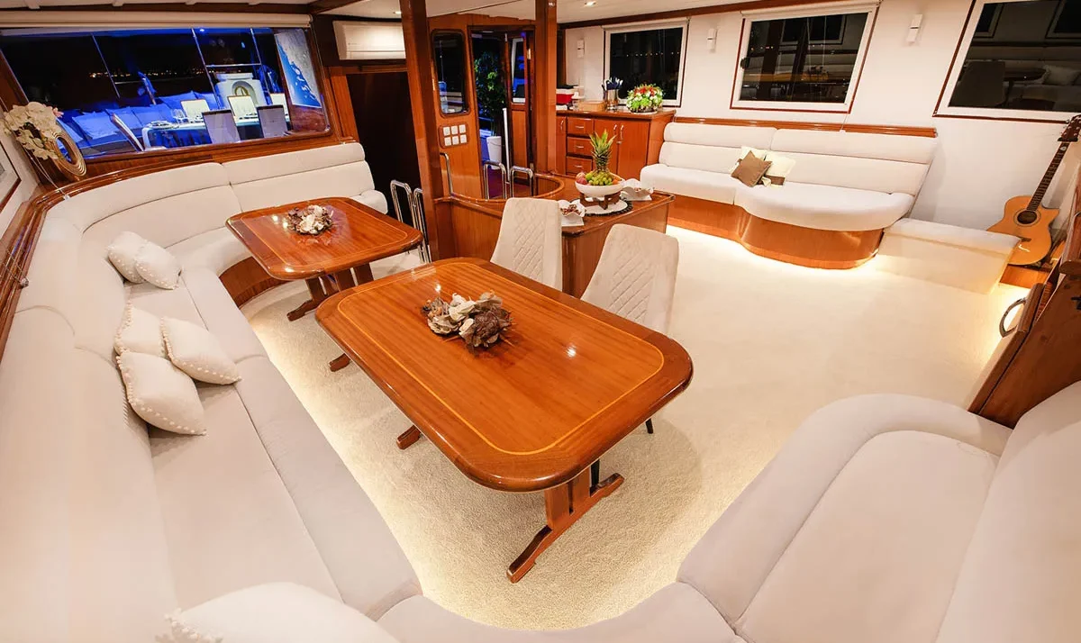 Luxury yacht salon with cream curved sofas, wooden dining tables, and a guitar resting by the window – warm, upscale interior.