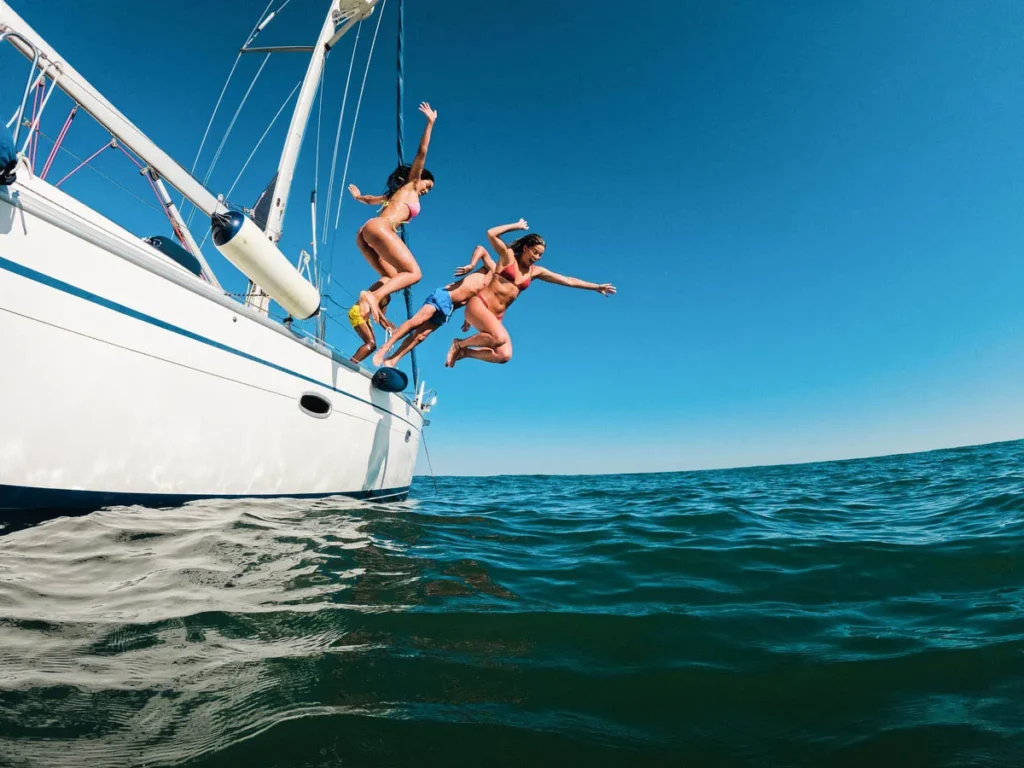 Group of Friends in Swimsuits Jumping from a White Sailboat into the Blue Ocean Under a Clear Sky