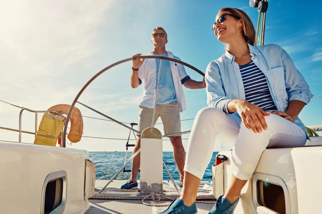 a Couple Sails on a Sunny Boat Woman Wearing Sunglasses and a Striped Shirt Sits at the Front Smiling As the Man Steers at the Helm with the Sea and Blue Sky Behind Them
