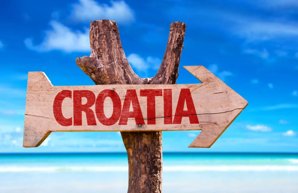 Wooden Sign with Red'CROATIA' text on a beach, blue sky and turquoise sea behind.
