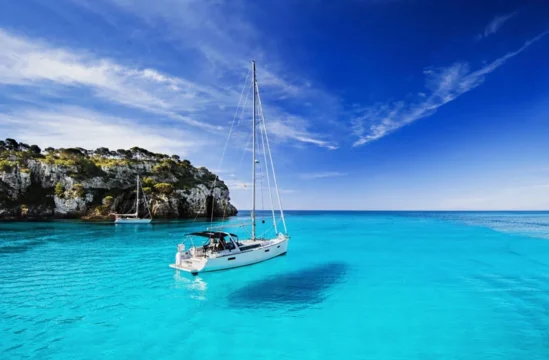 Sailboat Anchored in Bright Turquoise Water Near a Rocky Tree covered Coastline Under a Clear Blue Sky