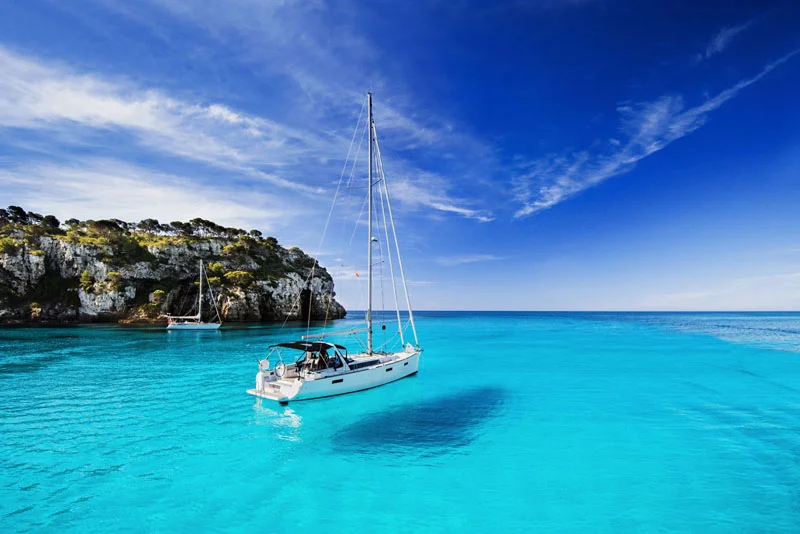Sailboat Anchored in Bright Turquoise Water Near a Rocky Tree covered Coastline Under a Clear Blue Sky