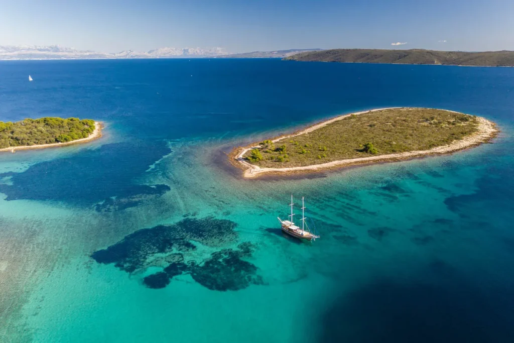 Aerial View of a Small Crescent shaped Island in Clear Blue Water with a Sailboat Anchored Nearby on Calm Seas