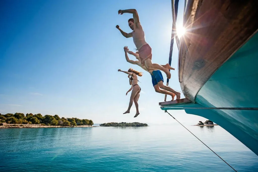 Group of Four People Jumping off a Turquoise Boat into Calm Blue Water on a Sunny Day Near a Tree lined Shore