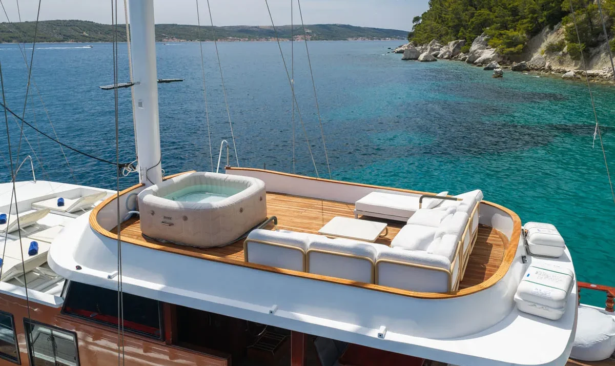 Luxurious yacht deck with white cushioned seating and a small hot tub, overlooking turquoise sea and rocky, forested coastline.