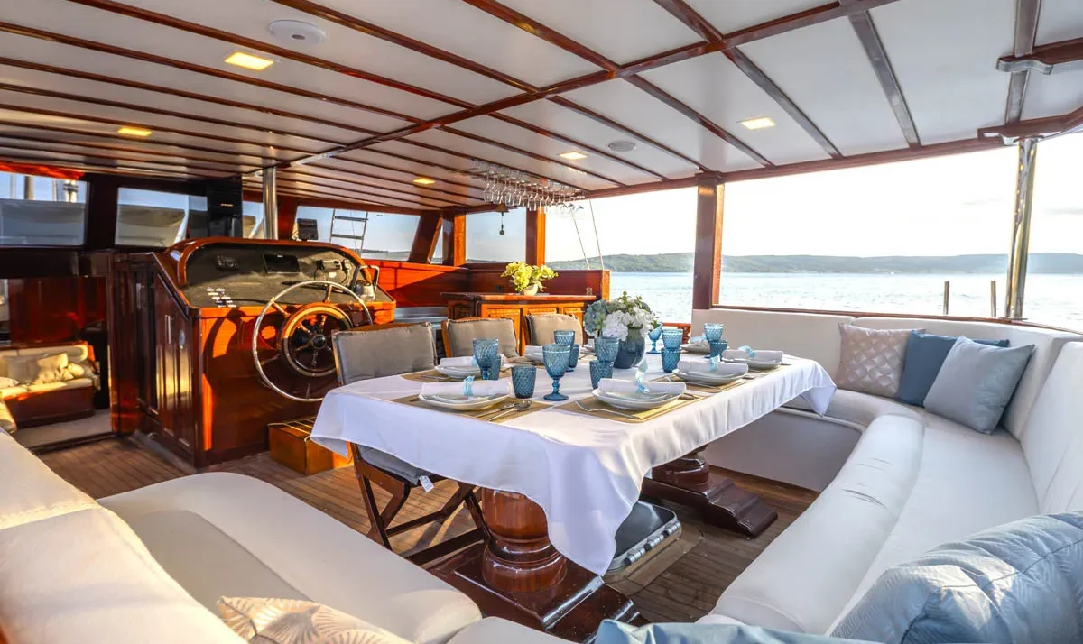 Luxurious yacht dining area: a table set with white cloth, plates, blue goblets, and a flower centerpiece, surrounded by cream seating and a sea view in the background.
