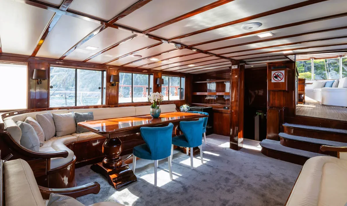 Luxurious yacht salon with wood-paneled walls, a curved beige sofa, blue chairs, and a glossy dining table by large windows.