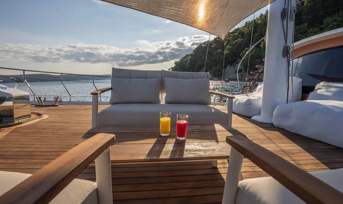 Luxurious yacht deck lounge with a wooden coffee table, two cushioned chairs, and two fruit drinks (orange and red) under a shade sail, overlooking calm water and a tree-covered coastline.