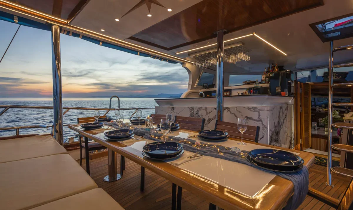Elegant yacht gulet Saint Luca dining table set for six with navy plates and wine glasses, overlooking a sunset over the sea.
