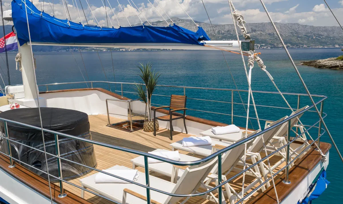 Deck of a sailing yacht with white lounge chairs, towels, a potted plant, and a covered table, overlooking calm blue water and distant coastline.