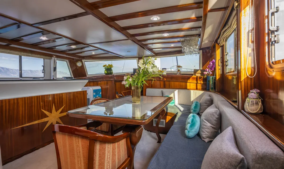 Luxurious yacht cabin with a glass-top dining table, gray corner sofa with teal and gray cushions, and dark wood paneling along the walls.