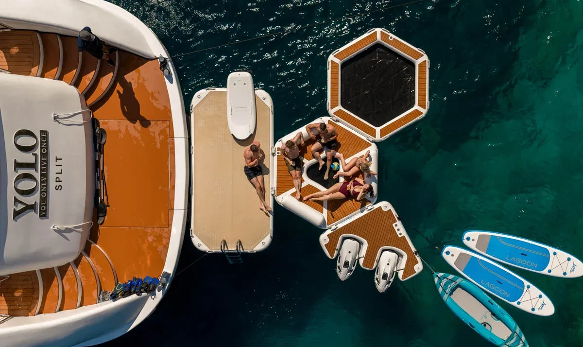 Aerial view of a yacht Yolo with orange deck and a floating platform where people sunbathe, with two blue Lagoon paddleboards nearby in turquoise water.