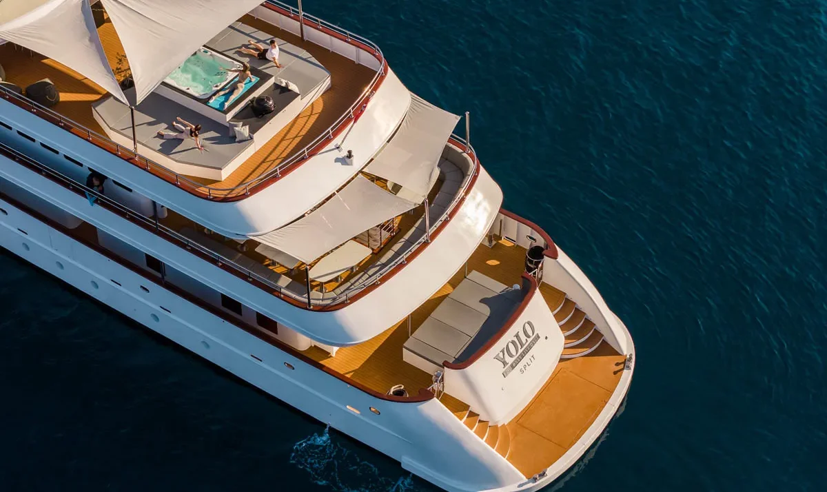Aerial view of a white luxury yacht with multiple decks and sun loungers, people relaxing on the upper deck, turquoise sea around it.