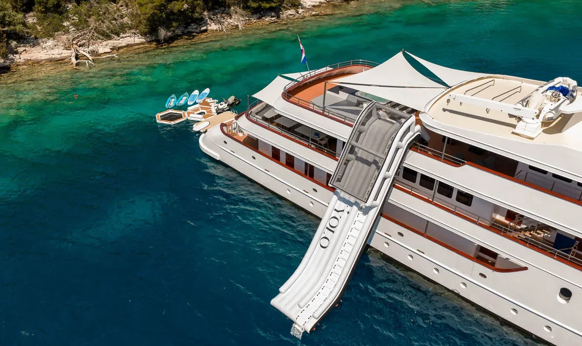 Large white luxury yacht with a tall waterslide extending from the deck into clear blue water near a rocky shoreline with paddleboards nearby.