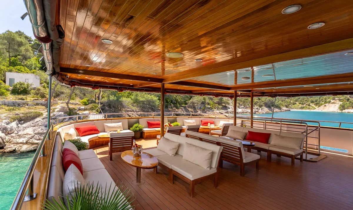 Open-air yacht deck lounge with beige cushions and red pillows, overlooking turquoise water and rocky coastline.