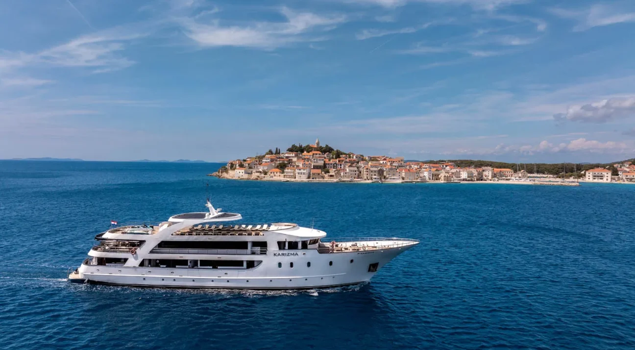 Large white luxury Mini cruiser Karizma sailing on calm blue sea in Croatia with a coastal town of red-roofed houses in the distance.