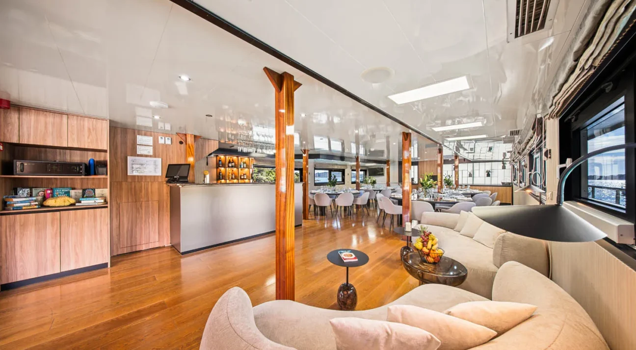 Luxurious, open-plan lounge on a yacht Karizma with beige curved sofas, wooden floors, a bar, and dining tables under a glossy ceiling.