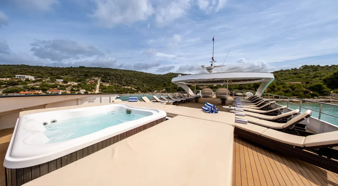 Luxurious yacht Karizma deck with a built-in hot tub, beige lounge chairs, and a shaded seating area overlooking a coastal landscape.