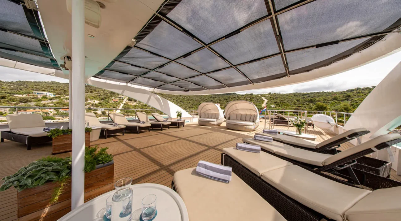 Luxurious yacht Karizma deck with beige loungers under a shaded canopy, potted plants, and a hillside view in the distance.