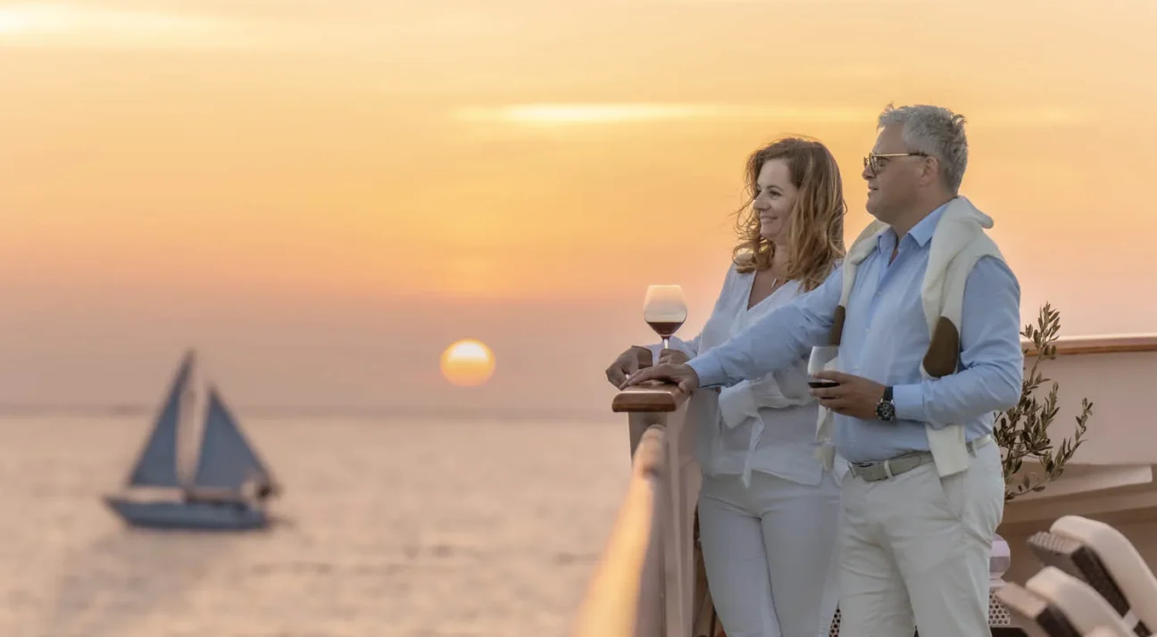 Couple standing at a yacht railing during sunset, holding wine glasses and smiling at the sea. A sailboat drifts in the calm water behind them.