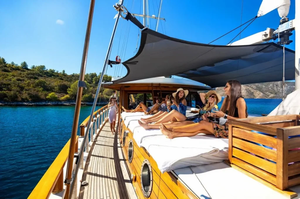 Group of Friends Sunbathing on a Wooden Catamaran Deck Under a Shaded Canopy Turquoise Sea and Rocky Coastline in the Background