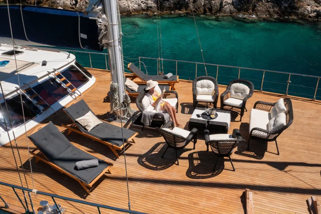 Person Relaxing on a Wicker Chair on a Wooden Yacht Deck with Turquoise Water Nearby Surrounded by Lounge Chairs and a Table Room for Cocktails