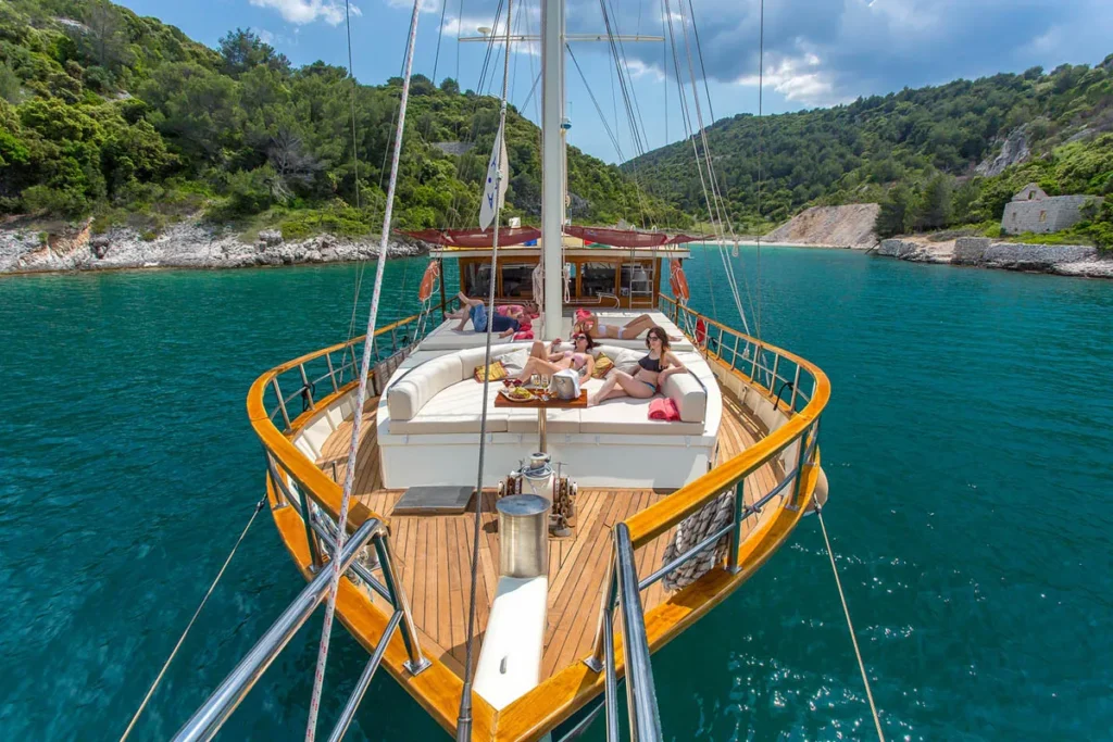 People Relaxing on a Wooden decked Sailing Yacht Sunbathing and Chatting As It Sits in Turquoise Water Near a Rocky Forested Shoreline