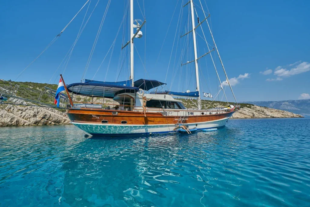 a Wooden Sailboat Anchored Near a Rocky Shoreline on Clear Blue Water Two People Jump into the Sea from the Side of the Boat Midday Sun