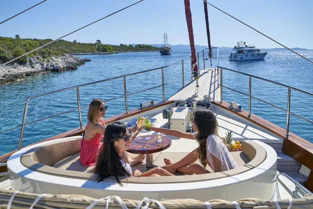 Three Women Toast with Cocktails on the Circular Lounge of a Luxurious Yacht Blue Water and Rocky Shoreline in View