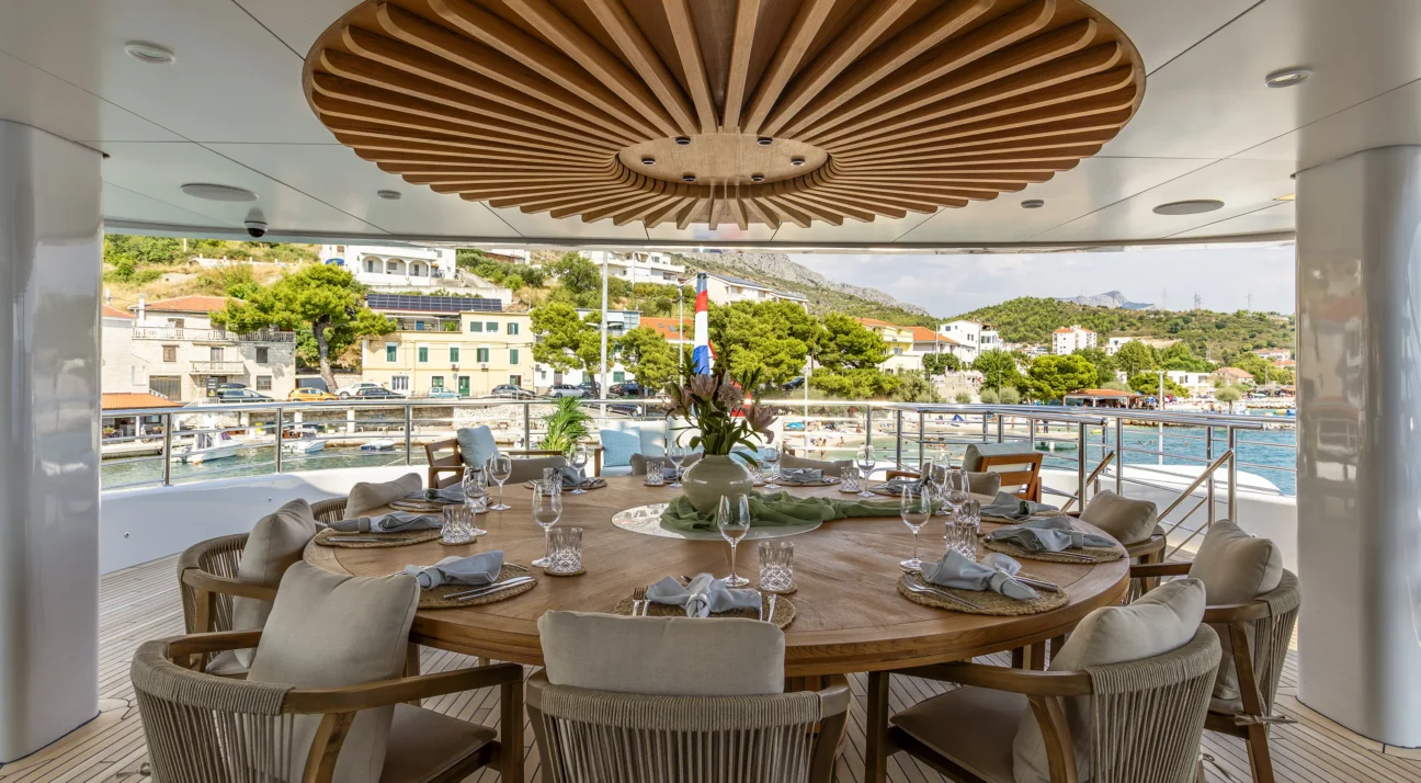 Round dining table on a yacht deck, set with glasses and napkins under a circular wooden ceiling centerpiece.