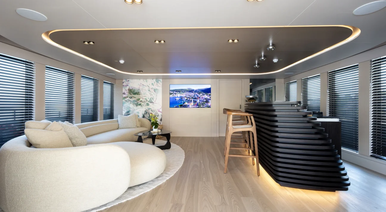 Modern living room with a large curved beige sofa, glass coffee table, and light wood floor; right side features a black layered sculptural bar or divider and stools.