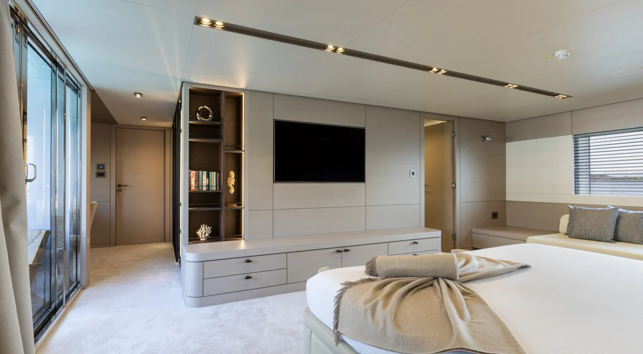 Modern luxury bedroom on a yacht with built-in beige cabinetry, a wall-mounted TV, and a plush bed topped with a beige throw blanket and pillows
