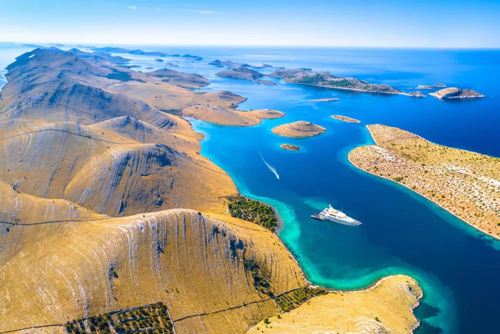 Aerial View of Arid Hills and Bright Blue Sea with a White Yacht Anchored in a Turquoise Inlet Far from Shore
