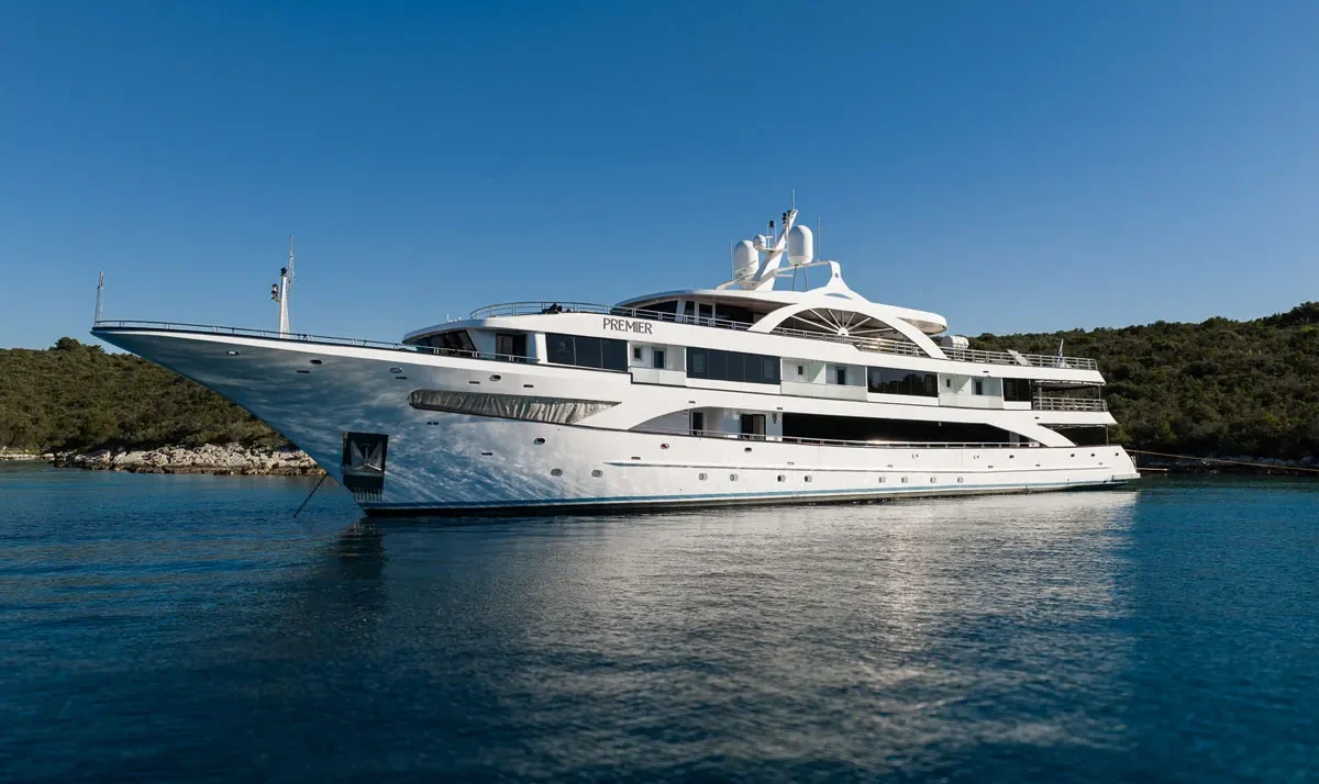 White luxury motor yacht named PREMIER anchored in calm blue water near a rocky coastline