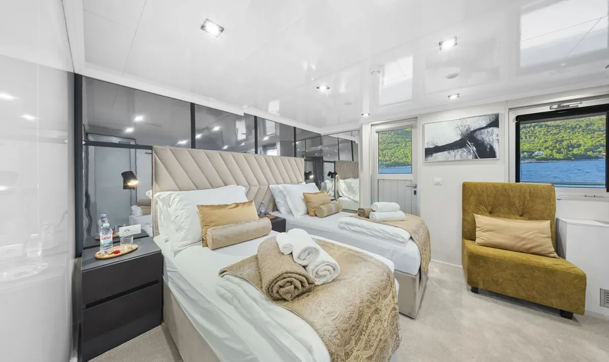 Luxurious yacht cabin with two twin beds, beige headboard, gold throw pillows, towels, and a water view through the windows.