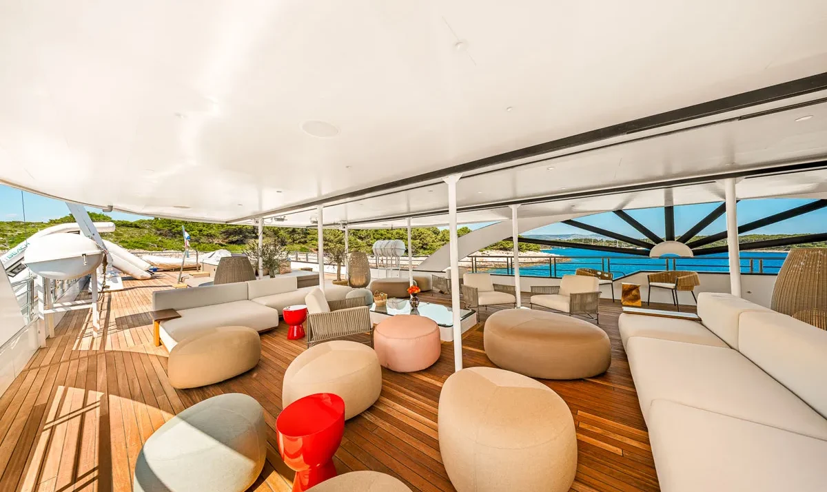 Luxurious yacht deck lounge with white cushioned seating, beige ottomans, and red side tables on a wooden floor, ocean view ahead.