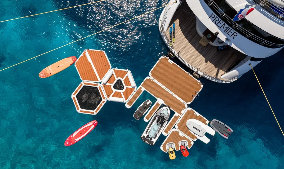 Aerial view of a luxury yacht with a modular wooden deck extending into blue water, with jet skis and paddleboards nearby.
