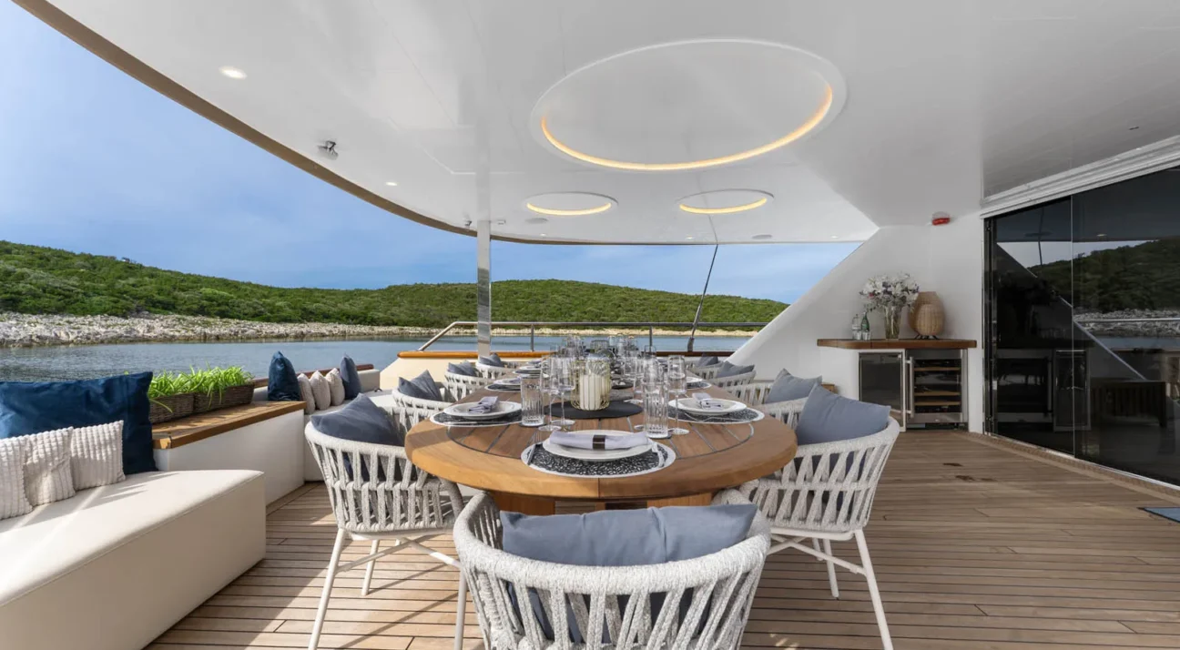Open-air yacht deck dining area with a long wooden table set for sixteen, wicker chairs, and an ocean shoreline in the background.