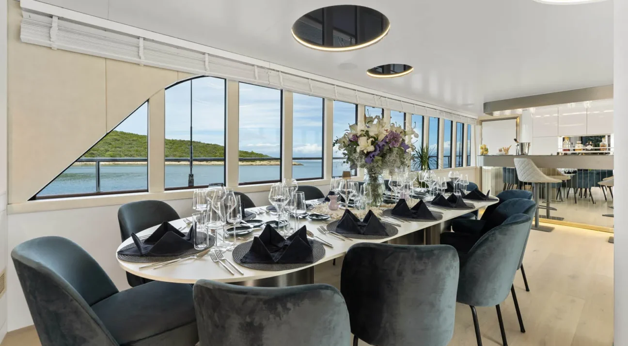 Formal seaside dining room with a long white table set for twelve, black napkins, glassware, and a floral centerpiece, surrounded by blue velvet chairs.