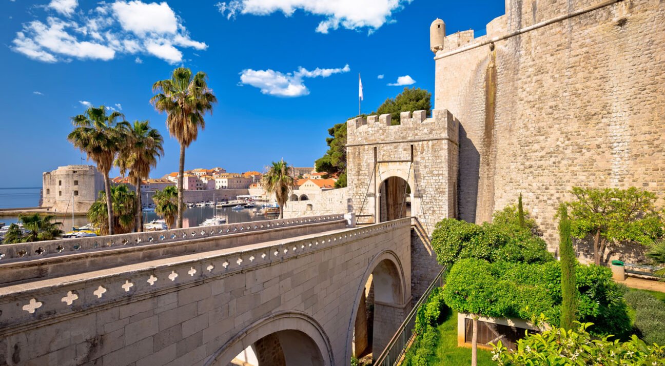 Stone fortress with crenellated walls, an arched bridge, and a marina of sailboats under a bright blue sky.