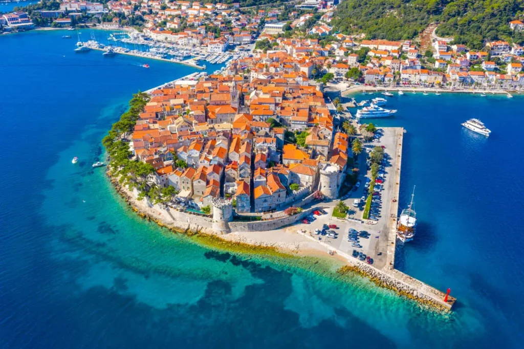 Aerial view of a walled medieval town with orange-tiled roofs on a rocky isthmus, surrounded by turquoise sea and a harbor with moored boats.",