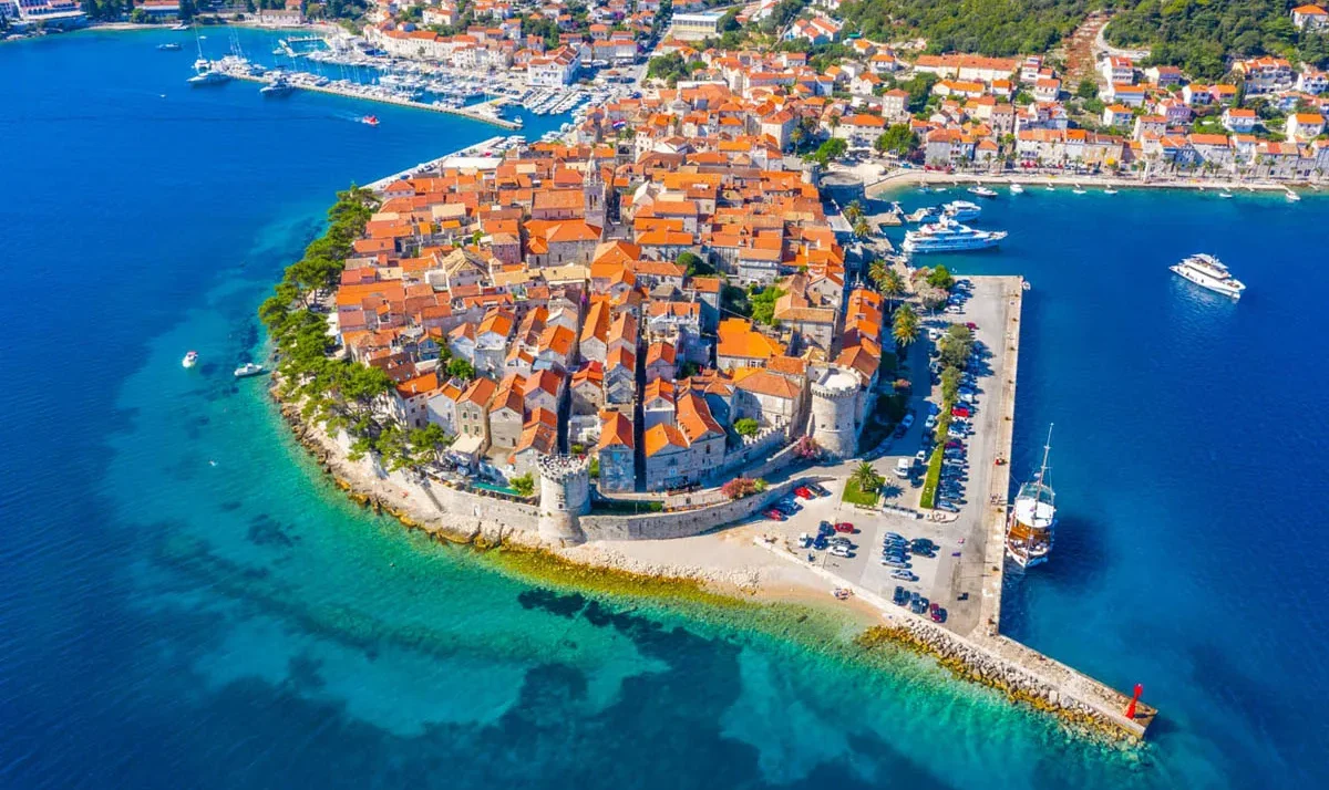 Aerial view of a walled medieval town with orange-tiled roofs on a rocky isthmus, surrounded by turquoise sea and a harbor with moored boats.",